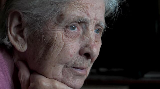 Close up profile of thoughtful old woman with grey hair and blue eyes, her face etched with wrinkles, looking away against a dark background, lost in contemplation and misses loved ones