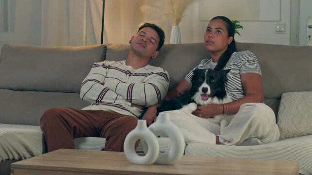 Young man falling asleep on a comfortable sofa while his wife and their border collie dog continue watching television together in a cozy modern living room during a late evening