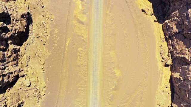 Overhead aerial view of a sandy desert road with tire tracks between rugged rock cliffs, ideal for travel, adventure, and remote landscape backgrounds.