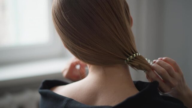 Close up of woman with long straight hair as woman ties it into low ponytail using her hands. scene is set indoors near window showing soft natural light and back of her neck and shoulders