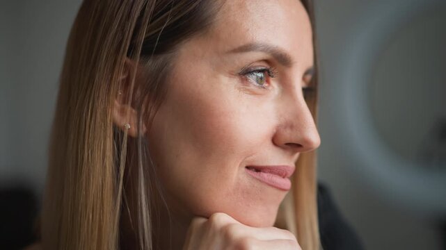 Close up of woman with long straight hair resting her chin on hand in soft lighting. Her expression is calm and thoughtful showing subtle facial features like lips and eyes. Background