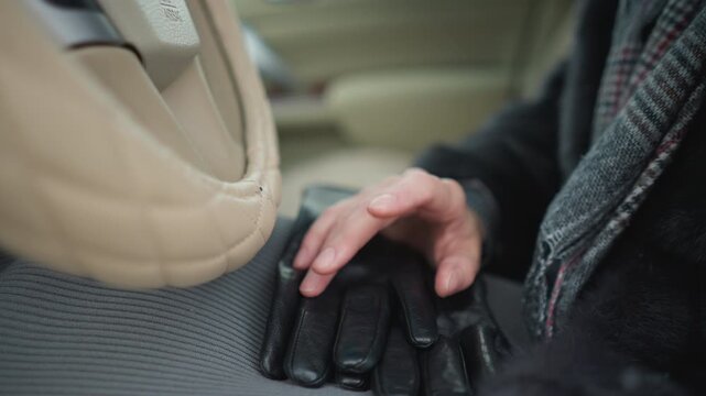 Close up of person hand resting on black leather gloves inside vehicle. person wears dark coat and patterned scarf with soft lighting highlighting texture of both hands and materials