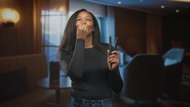 Young woman holds straightener and brings finger to lips to blow a kiss while smiling in a building lounge with chairs and lamp; self care playful.