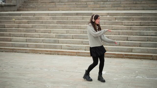 Close up of young woman dancing energetically in front of stone steps wearing casual winter clothing including headphones. Her movements are fluid and expressive against backdrop of large