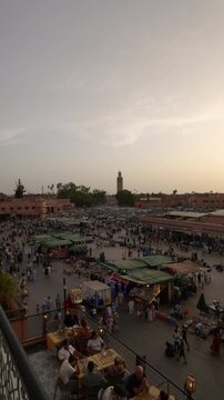 Marrakech, morocco. August 9, 2026. Crowded jemaa el fnaa square at dusk with the koutoubia mosque in the background. Tourists eating at a rooftop cafe overlooking the market