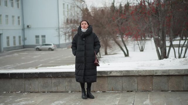 Close up of woman standing outdoors in winter wearing long black puffer coat and holding hands behind her back. scene includes snow covered ground bare trees with red berries parked cars