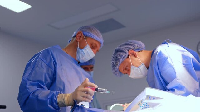 Neurosurgeon using tool with the tube stands leaned over the patient. Male colleague is holding a syringe filled with saline solution. Neurosurgical operation in progress.