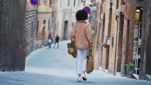 Woman with a summer hat strolling on street alley pavement in Siena, Tuscany, Italy