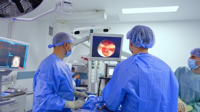 Team of doctors, standing near the operating table with the patient on, look attentively at the screen in front of them. Inside the modern well-equipped surgery room for neurosurgical operations.