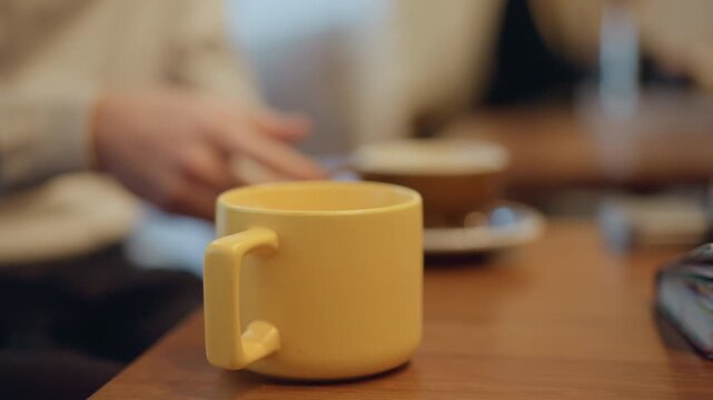 Close up of yellow ceramic mug on wooden table with soft lighting. Background shows blurred person and coffee cup suggesting casual indoor setting like cafe or home workspace