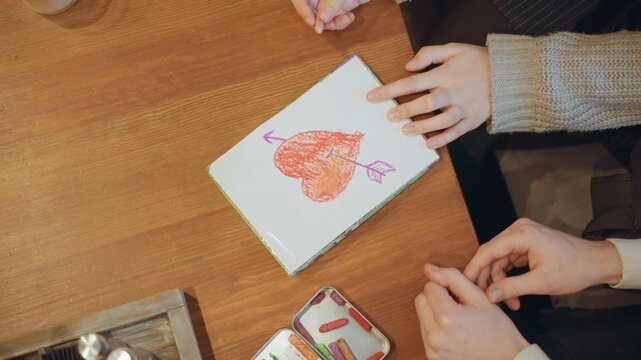 Close up of hands drawing heart with arrow on paper using pastels. woman hand in knitted sleeve and another woman bare hands work together at wooden table surrounded by crayon tins and soft