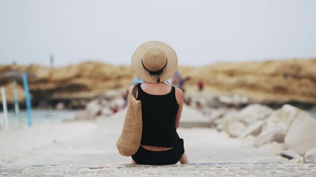 Back view of stylish woman in a hat sitting by the rocky beach in Monastir, Tunisia