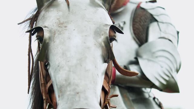 Closeup shot captures rain droplets on steel plate armor as a mounted knight in full harness sits on a brown warhorse wearing a steel shaffron face shield in rainy weather...