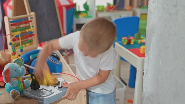 Boy presses a yellow toy drill with both hands on a playroom workbench amid toys and a blue telephone; playful curiosity.