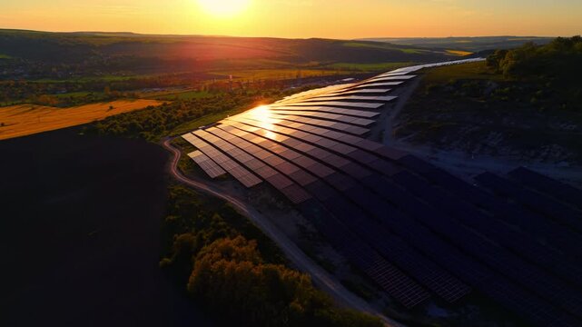 Elevated view of solar farm at sunset, hillside. Above shot of panel rows descending toward a village nestled in the valley below.