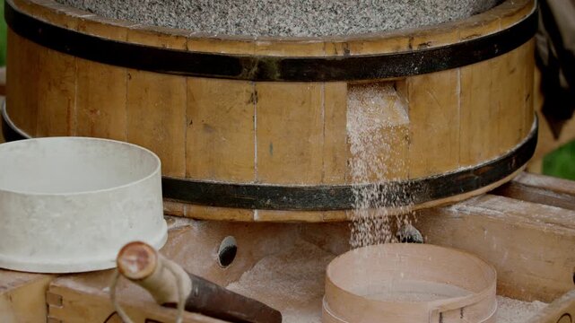 Closeup of traditional medieval rotary stone hand mill with granite millstones grinding wheat grain into fresh coarse wholemeal flour falling from wooden spout into round sieve...