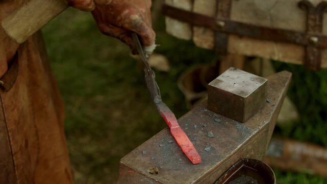 Detailed view shows blacksmith artisan striking glowing hot orange metal with steel hammer to shape blade or tool while scale dust flies on rustic anvil surface...