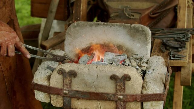 Medium shot captures medieval blacksmith holding iron tongs to heat metal workpiece in a traditional portable stone hearth filled with burning charcoal and glowing fire.