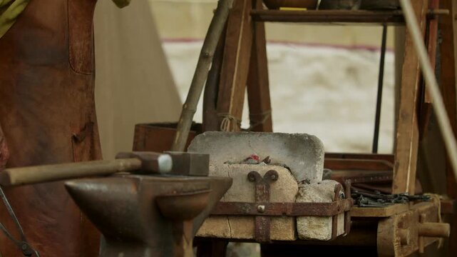 Closeup of medieval blacksmith stirring burning coals with long tongs to heat a red-hot iron piece in a stone forge while pumping air manually with a wooden lever...