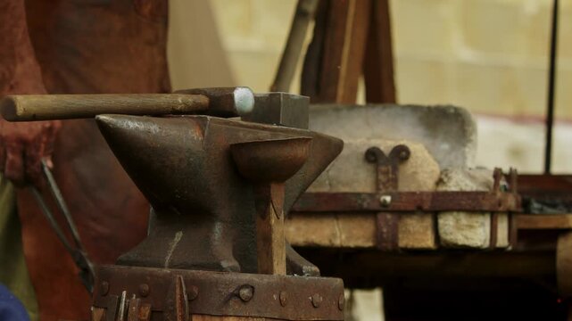 Closeup of a heavy iron anvil with a forging hammer resting on top while a medieval blacksmith works the wooden bellows lever to stoke the stone forge in the background...