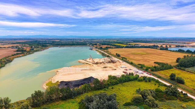 Aerial view of a sand quarry near turquoise lake. Industrial processing equipment and sand piles sit along the shore under a partly cloudy summer sky.