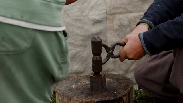 Young apprentice hands grip blacksmith tongs and wooden mallet to strike a steel die tool on a tree stump anvil during an interactive medieval market workshop...