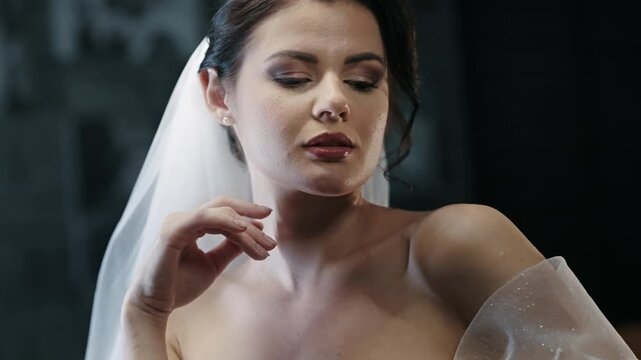 Seductive bride wearing a delicate veil poses elegantly looking in camera with teasing look in a dimly lit setting