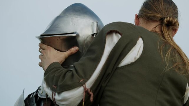 Closeup shot shows medieval squire carefully adjusting, fitting, and securing polished steel frog-mouth helmet onto knight's head before tournament battle.