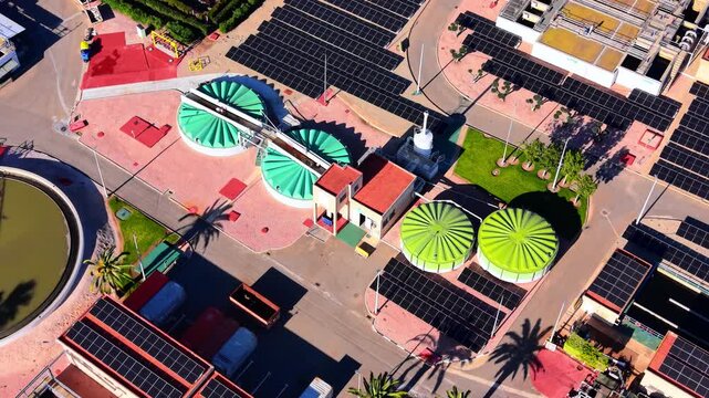 Overhead view of biogas digesters at a treatment plant, Spain. Green domed tanks and solar panels sit beside farmland under bright sunlight.