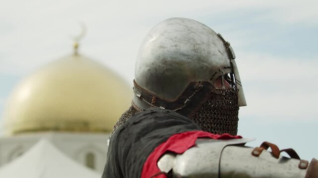 Dramatic close view focuses on crusader warrior in iron helmet and mail coif overlooking golden Islamic dome and minaret during historical crusade reenactment.