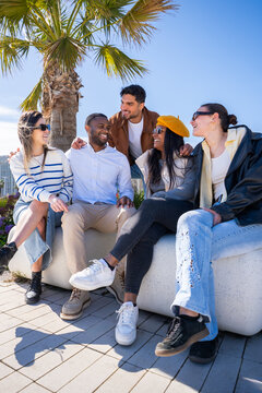 Vertical. Multicultural group of friends sit together on a bench outdoors in a bright residential district, chatting and enjoying leisure time under palm trees.