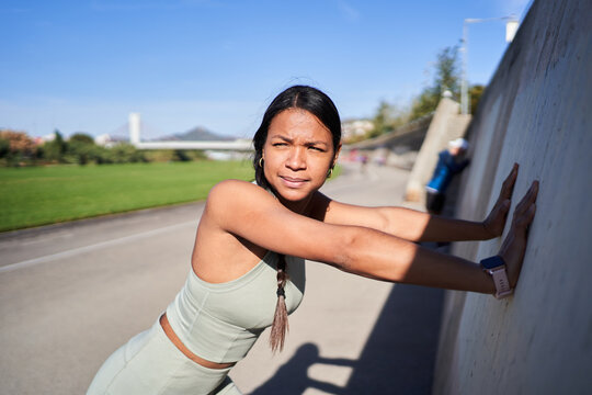 Focused Black Latina athlete stretching her upper body against a concrete wall during an outdoor workout. Sunny fitness scene conveying preparation, flexibility and recovery.