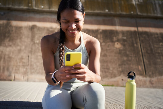 Smiling Black Latina athlete using her smartphone while sitting outdoors during a workout break, with a reusable water bottle beside her in bright sunlight.