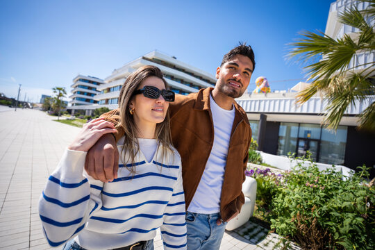Stylish young couple stroll together outdoors in a clean modern residential district with palm trees and bright sunshine, enjoying a relaxed lifestyle moment.