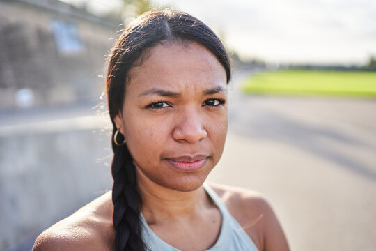 Confident close-up portrait of an African millennial woman standing outdoors in park after exercise, wearing sportswear and looking at camera. Natural light portrait shows strength active lifestyle