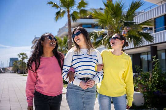 Multicultural female friends walk side by side and smile in a sunny residential district, sharing a casual friendship and lifestyle moment.