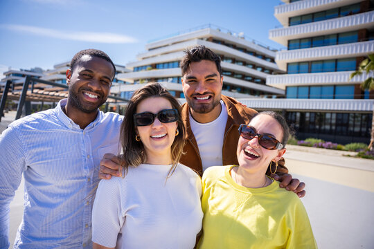 Multicultural group of friends pose together and smile at camera in a bright urban residential setting with contemporary buildings and blue sky.