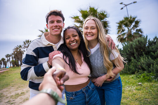 Three diverse young friends smile while holding the viewer's hand and leading them forward through a sunny green park, conveying friendship and shared adventure.