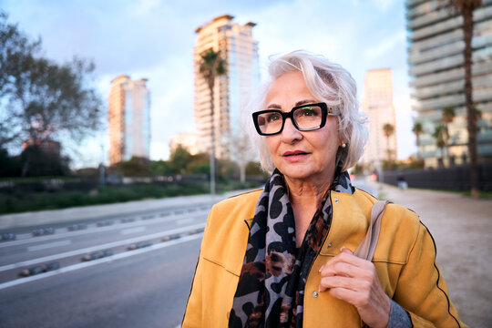 Elegant senior woman serious face with stylish glasses and a yellow jacket walks confidently in a modern city at dusk, enjoying her retirement and urban lifestyle