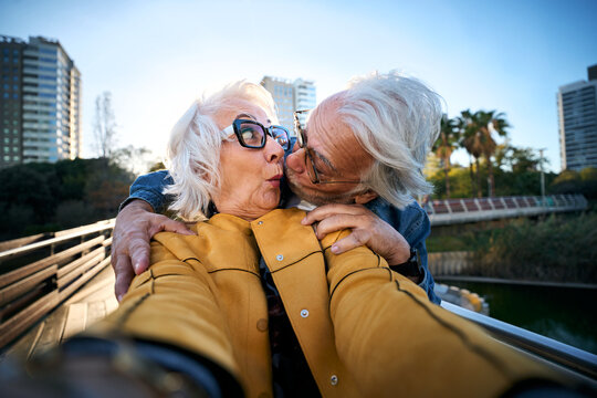 Playful senior couple taking a close selfie as the man kisses the surprised woman on the cheek. Wide-angle urban scene conveying affection, humor, companionship and active aging.