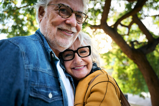 Close up of laughing mature adult Caucasian couple looking at the camera standing embraced affectionately in a park. Elder people in love enjoying romantic time. Positive pensioner relationships