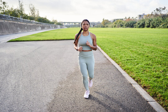 Full body african millennial woman jogging outdoors in park on sunny day, wearing sportswear and smartwatch. Expression shows energy, discipline and determination in active lifestyle.