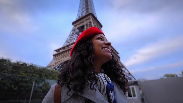 African millennial smiles beside the Eiffel Tower in Paris at blue hour, wearing a red beret and coat, capturing urban travel vibes with direct gaze.