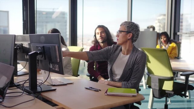 Mature businesswoman calling a colleague over to her desk to ask a question while other employees work nearby. Candid office scene showing communication, coordination and teamwork.