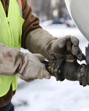 Winter road worker in high visibility vest adjusts salt truck valve with insulated gloves in bright snowy daylight, close cropped view showing careful maintenance work and cold weather conditions