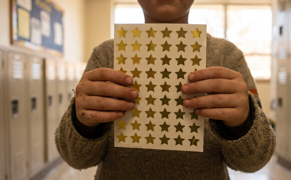 Young schoolboy holding a sheet of gold star stickers in a school hallway with lockers
