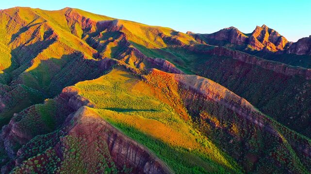 Sunlit aerial panorama of rugged, layered mountains and green slopes, creating a dramatic nature travel background.