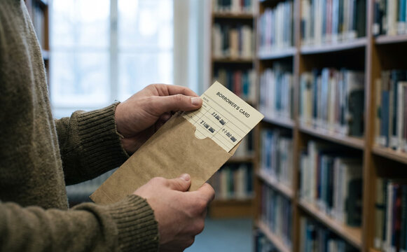 Young adult man holding library book pocket with borrower card in a university library stack