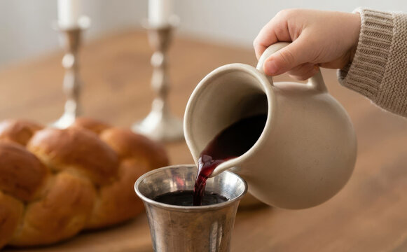 Child pouring red wine from a ceramic pitcher into a silver cup next to challah bread for Shabbat dinner