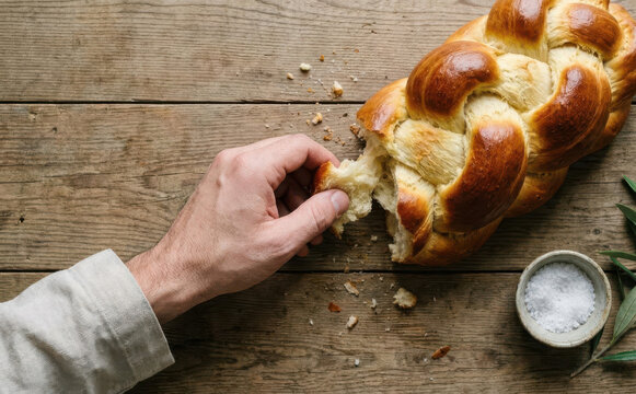 A male hand tearing a piece of freshly baked braided challah bread on a rustic wooden table